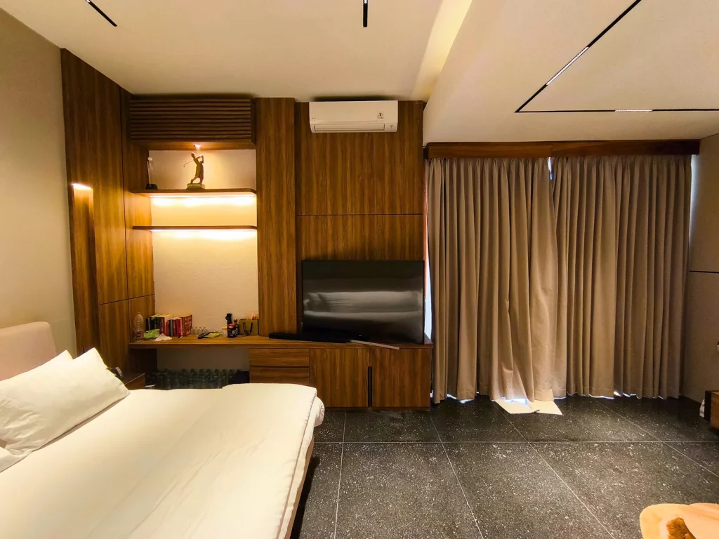 Bedroom media and workspace wall in a Kerobokan master suite, Bali, with walnut joinery, backlit display shelves, an integrated desk and full-height curtains, designed by Bali Best Design